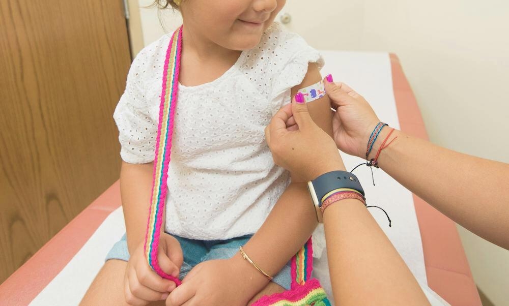 Child sitting on a medical examination table receives a colorful adhesive bandage on the arm from an adult, in a clinic room with wooden door and white walls.