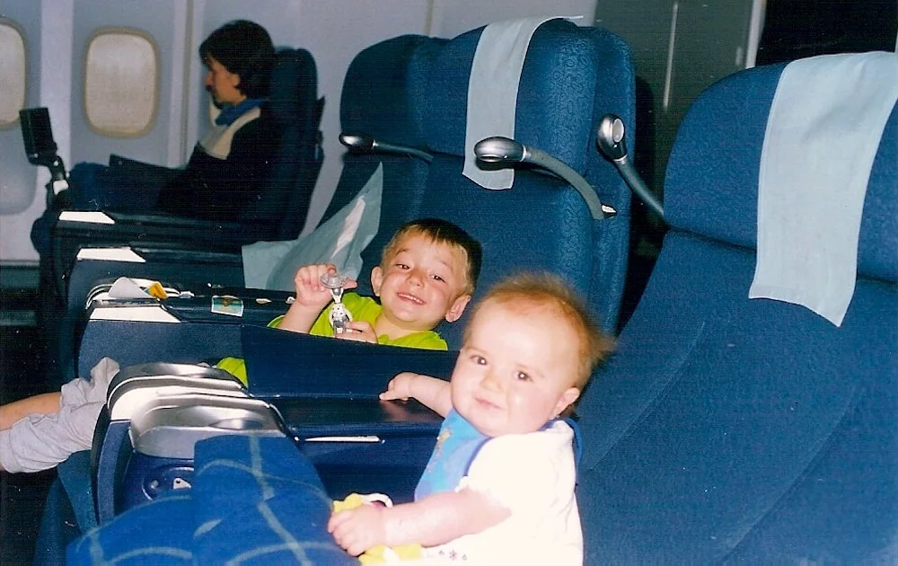 Two young children sit in airplane seats, one holding a toy and smiling, the other looking at the camera. Behind them, a person sits quietly, reading in a row of blue seats.