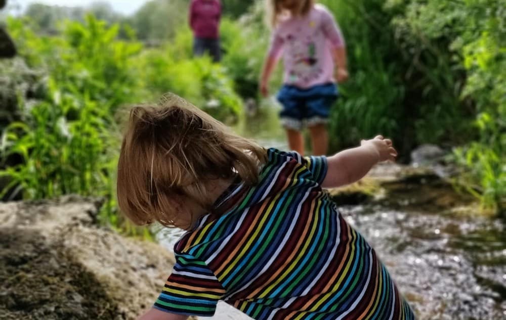 A child in a striped shirt reaches over a rock in a lush, green outdoor setting with two other people in the background near a stream.