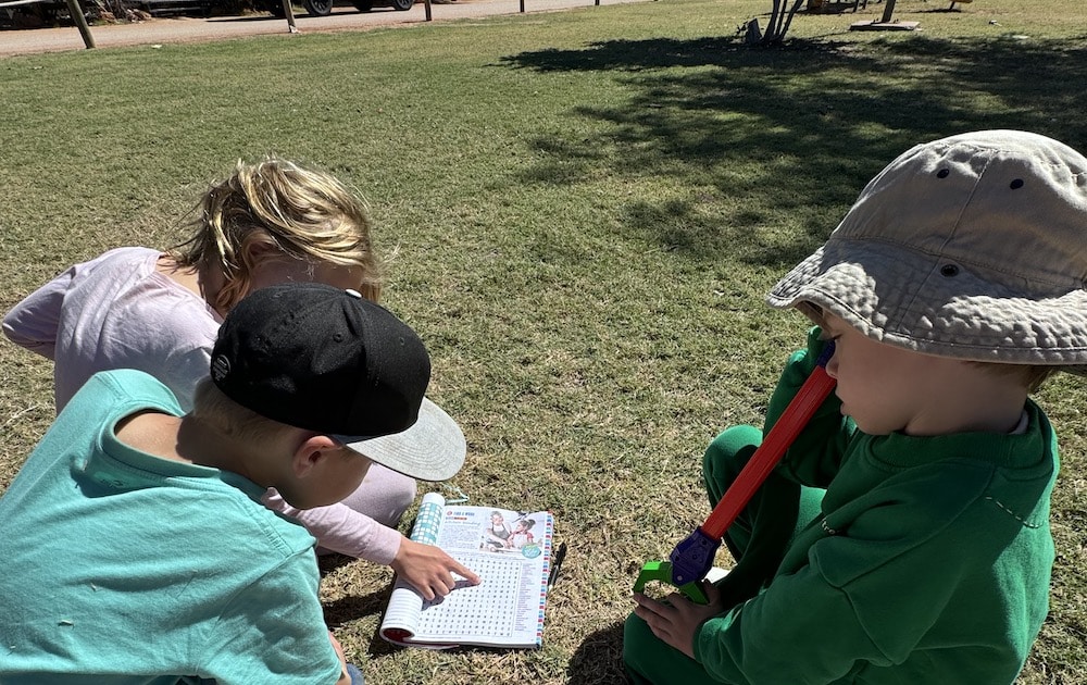Children are sitting on grass, reading a colorful book together on a sunny day. One child wears a sun hat, holding a toy, adding a playful touch to the scene.
