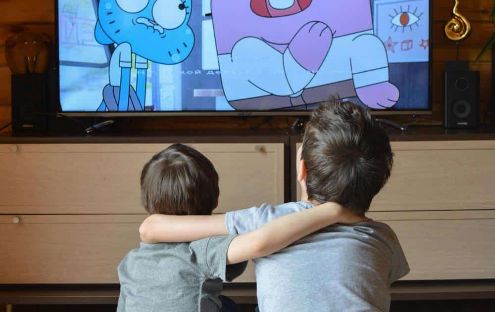 Two children watch cartoons on a television, sitting on a floor with one child's arm around the other. They are in a cozy room with wooden accents and a dresser.