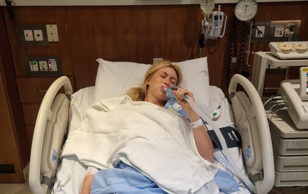 A woman is lying in a hospital bed, using a breathing mask. The room is equipped with medical devices, including monitors and emergency buttons, suggesting a clinical setting.