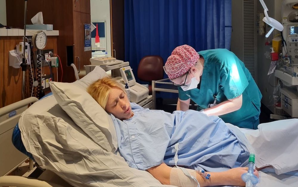 A woman in a hospital bed appears to be in labor while a medical professional, wearing scrubs and a mask, attends to her. Medical equipment surrounds the hospital room.