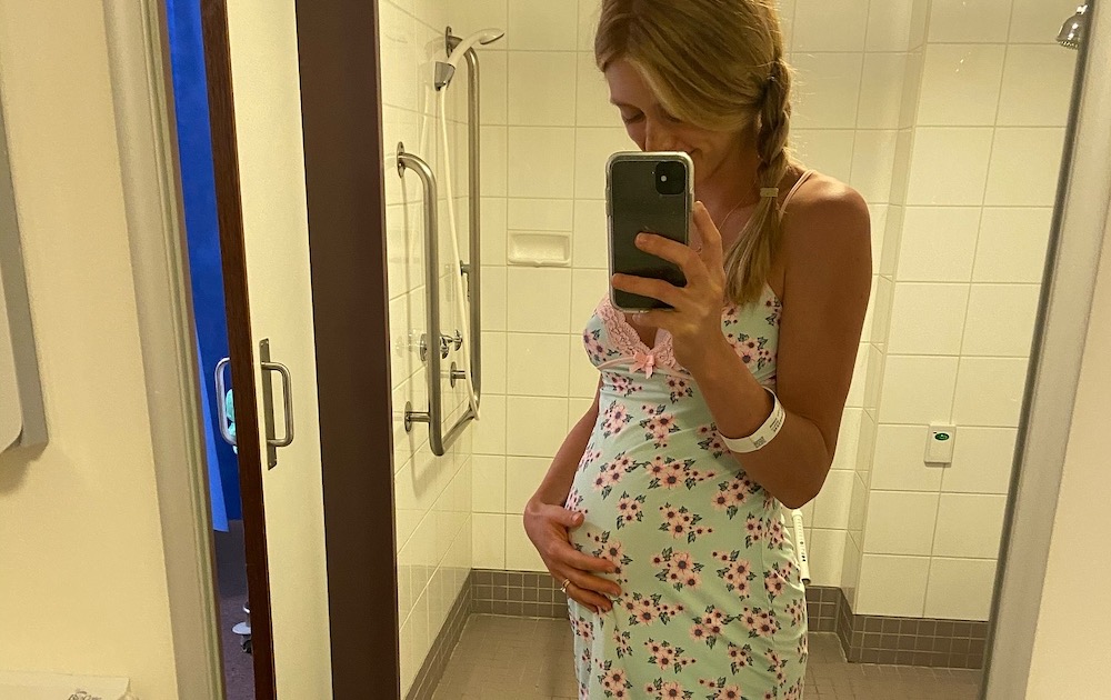 A woman in a floral dress takes a mirror selfie, cradling her belly in a tiled bathroom, with handrails visible on the wall. She wears a wristband.